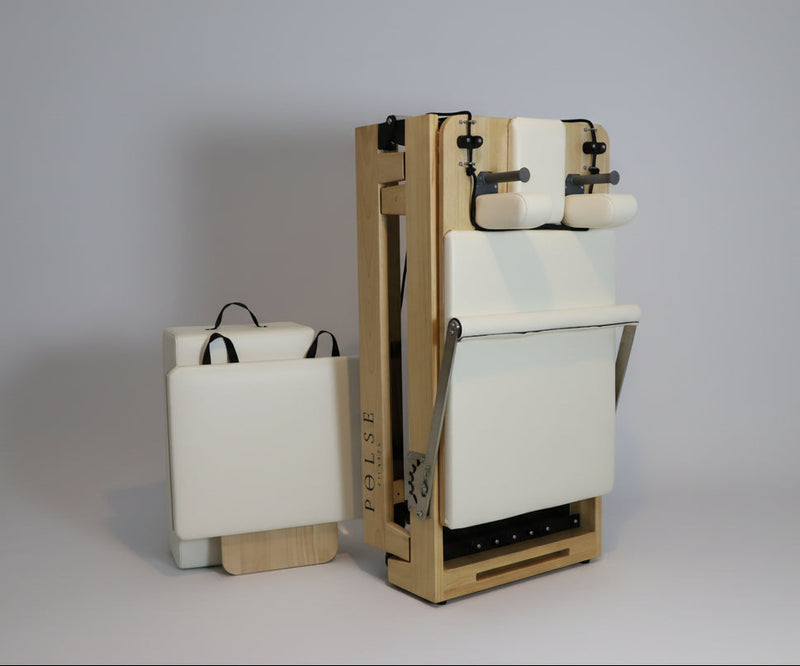 Foldable wooden Pilates reformer machine with cream upholstery shown in upright storage position beside matching carry case — ideal for small-space home workouts